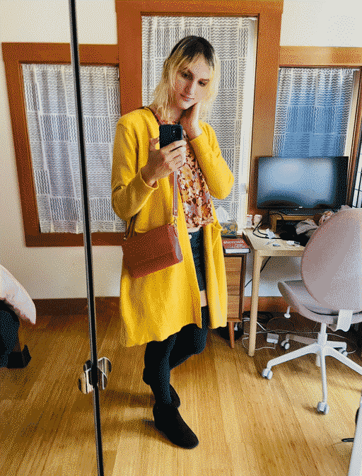 A full-body mirror selfie of me. I'm look affectionately at my phone with my hand on my face. I have long bleach blond hair that hasn't been bleached in half a year, which you can tell from the roots that nearly reach my eyebrows. I'm wearing a floral blouse, a denim miniskirt, and thigh-highs under a big comfy knee-length cardigan. I have a cute lil leather purse on my side and black suede high-heel boots. My tum shows just a little in the gap between my blouse and skirt.