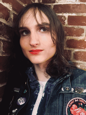 A chest-up selfie of me in front of a brick wall, looking into the distance. I’m wearing a denim jacket with pins and patches on it, one saying “Slowly but surely.”